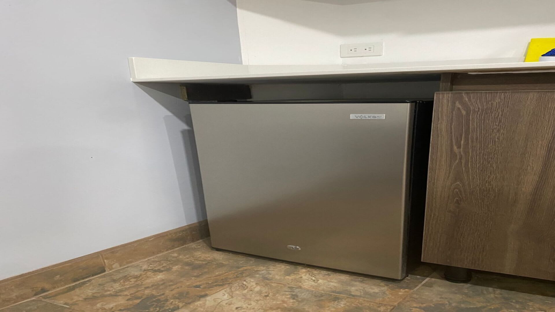 A compact refrigerator in a corner with a white countertop above and a wooden cabinet to the right.