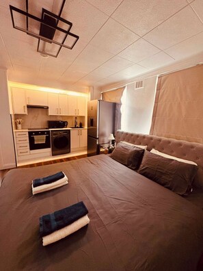 Een slaapkamer met een groot bed, een aparte kitchenette en een raam met gordijnen.