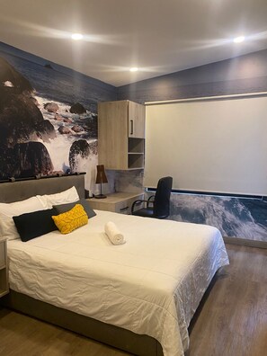 A bedroom with a bed, a desk, a chair, a wardrobe, and a large window with a view of the sea. - Laureles Executive Suites 202 (Medellín)