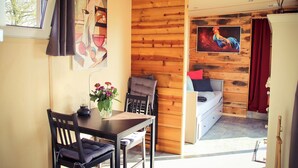 Une pièce avec un mur en bois, un lit avec un rideau rouge, un tableau représentant un coq, une table à manger avec des chaises et une fenêtre donnant sur des arbres.