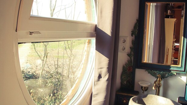 Une pièce avec une fenêtre ronde, un miroir, un lavabo et une chaise rouge.