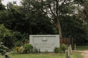 Señal de 'MUCHENJE River Lodge' con una puerta y follaje circundante.