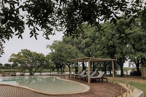 A wooden deck with a swimming pool, lounge chairs, and a pergola. - Muchenje River Lodge  (Katima Mulilo)