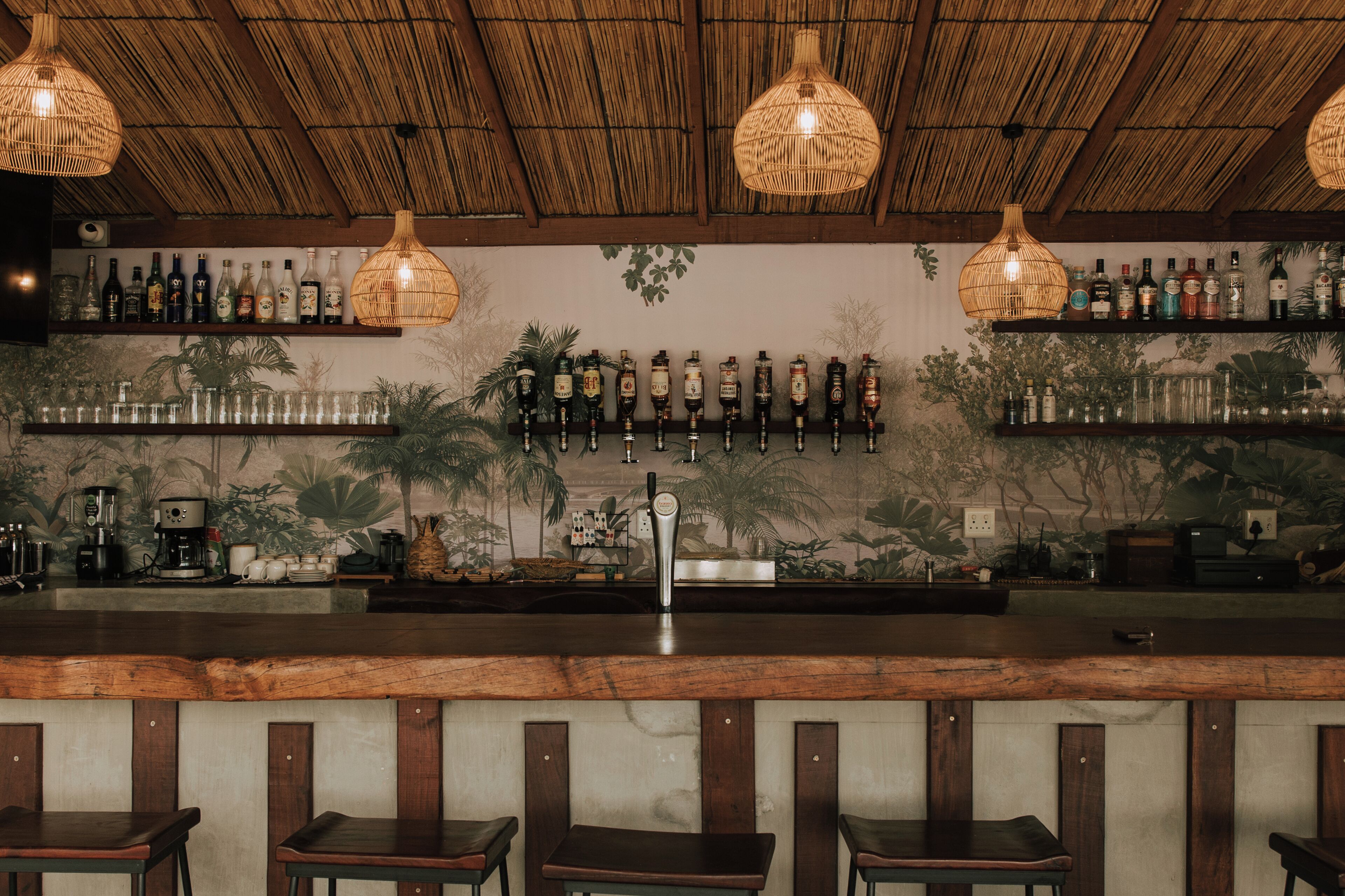 Zona de bar con mostrador de madera, taburetes, lámparas colgantes de mimbre y estantes con botellas.