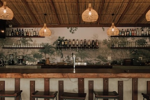 A bar area with a wooden counter, bar stools, hanging wicker light fixtures, and shelves with bottles. - Muchenje River Lodge  (Katima Mulilo)