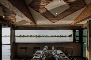 A dining area with a table set for four, large windows, and a view of a body of water. - Muchenje River Lodge  (Katima Mulilo)