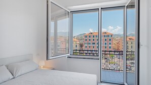 Ein Schlafzimmer mit Bett, Fenster und Blick auf Gebäude sowie ein Balkon mit Geländer.