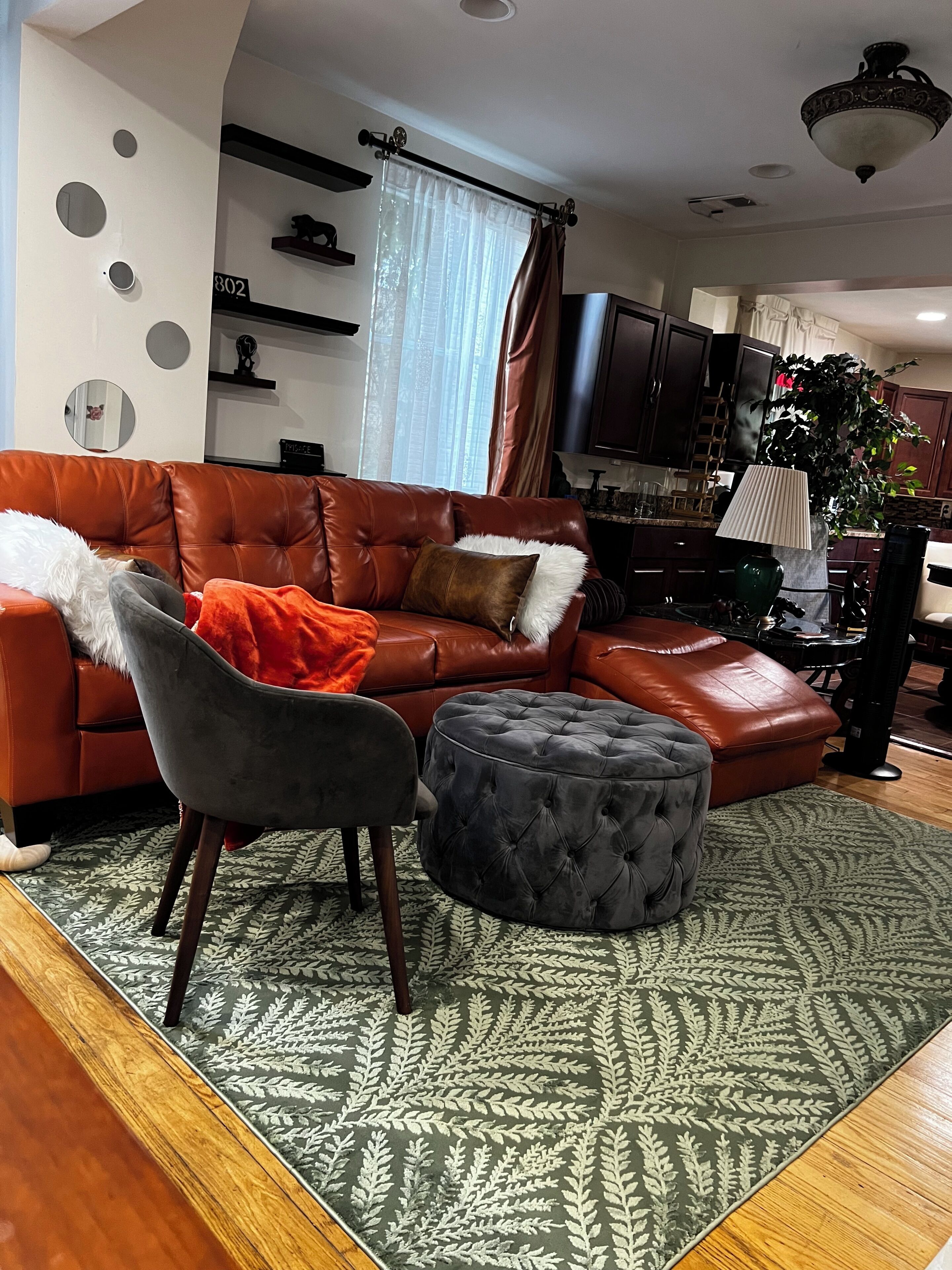 Un salon avec un canapé en cuir marron, un fauteuil gris et un pouf à motifs. Un tapis orné recouvre le sol.