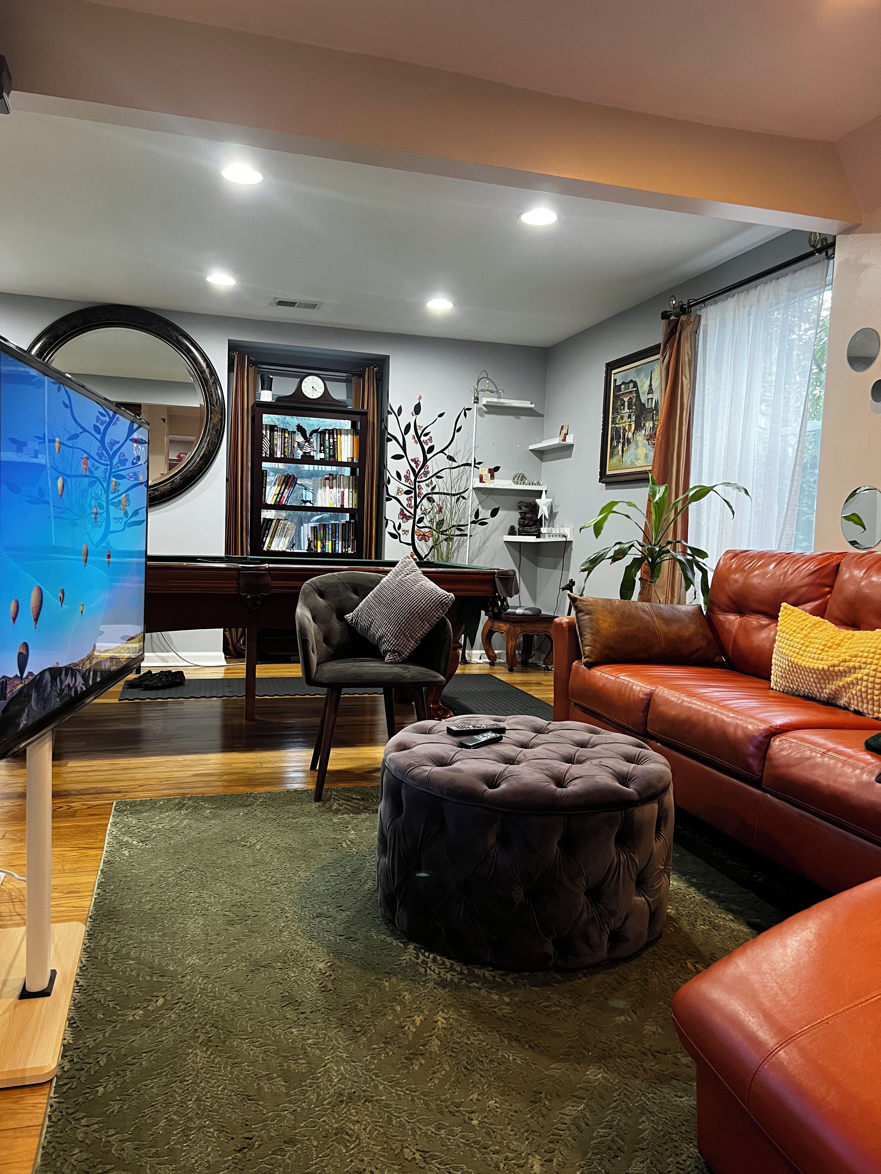 A living room with a leather sofa, a tufted ottoman, a bookshelf, a flat-screen TV, and a decorative mirror.
