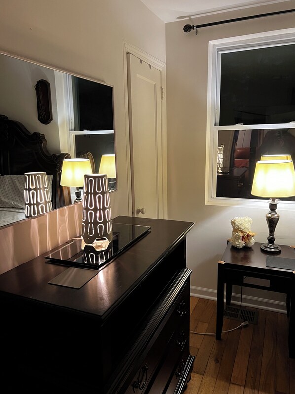 Une pièce avec une commode en bois foncé, un miroir, une table de chevet avec une lampe et une fenêtre.