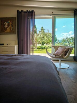 A bedroom with a large bed, a chair with a cushion, a painting, and a view of a garden through a glass door. - Villa among vineyards with pool near Conero (San Marcello)