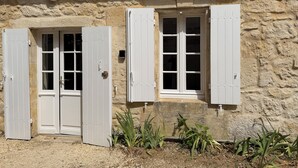 Ein Steinhaus mit zwei Fenstern mit weißen Rahmen und einer Tür, ein kleiner Garten mit Pflanzen sowie ein Metallfallrohr am Dach.