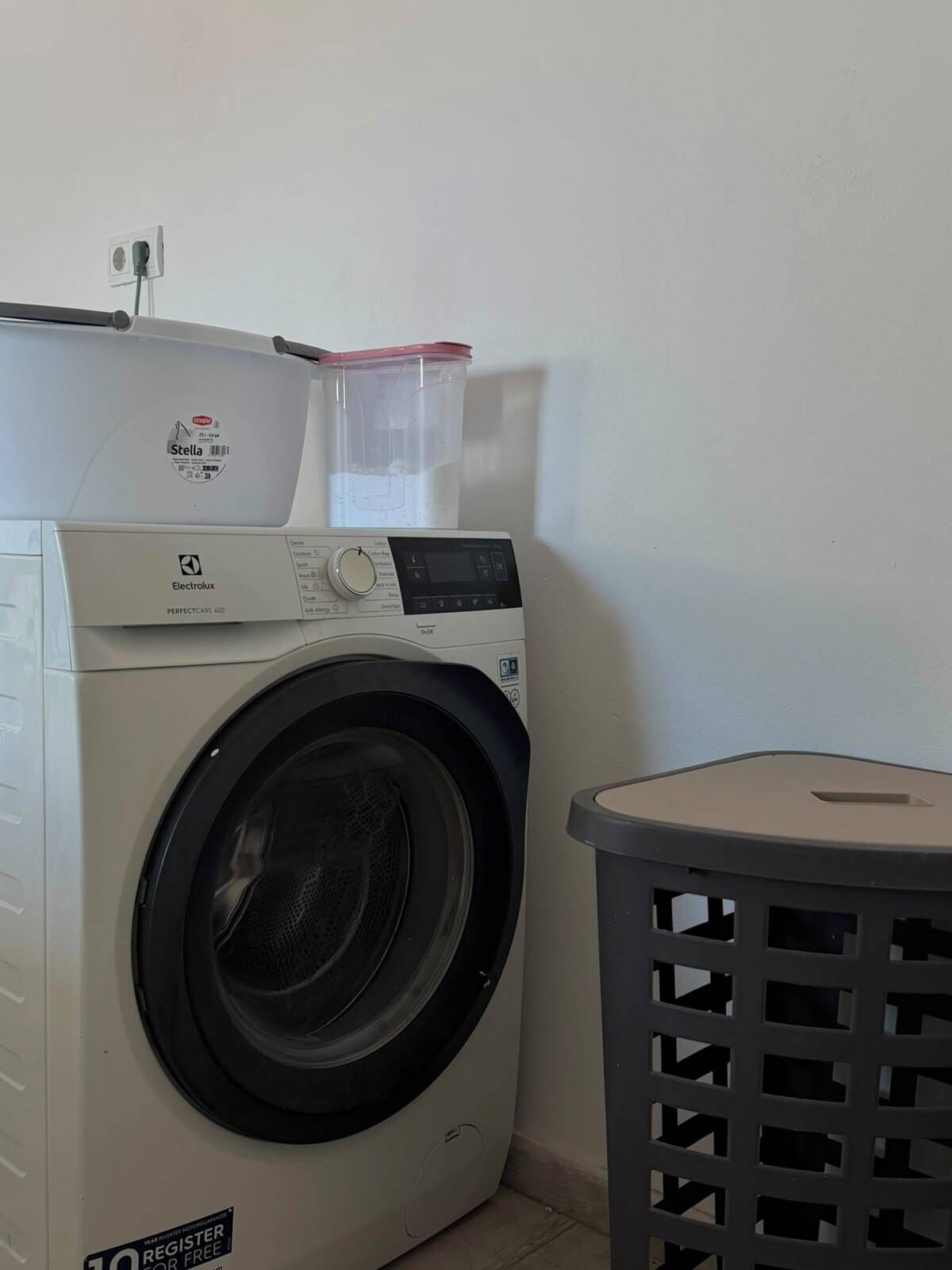 Una lavadora compacta con panel de control, un recipiente de plástico encima y una cesta de ropa a un lado.