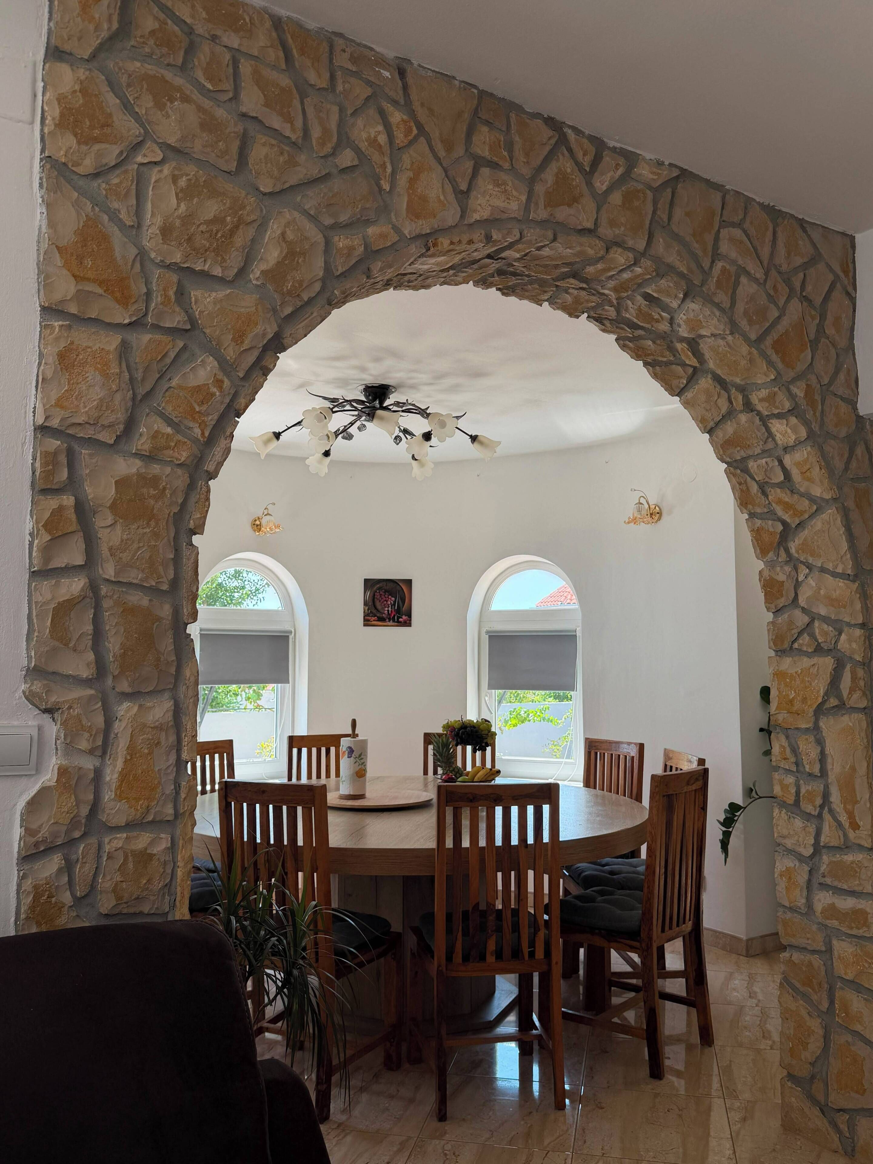A dining area with a stone archway, wooden chairs, a round table, and a chandelier.