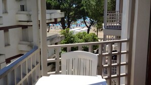 A balcony with a white table and chair, overlooking a beach with umbrellas and trees. - Spacious and central stay with sea views (Lignano Sabbiadoro)