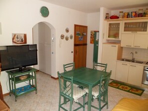 A living room with a TV, a dining table with chairs, a kitchen with cabinets and a sink, and a clock on the wall. - Practical stay in Lignano with parking (Lignano Sabbiadoro)