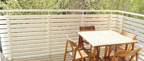 A wooden deck with a table and chairs, surrounded by white railings, overlooking a dense green forest.