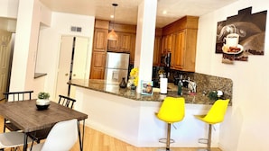 A modern kitchen with wooden cabinets, a stainless steel refrigerator, and a granite countertop. There are two yellow bar stools and a dining table with chairs. - Medical center 2-bedroom apartment in perfect Houston with cool AC and comfort (Houston)