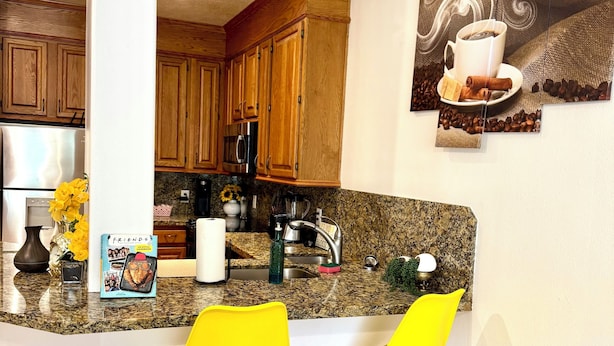 A kitchen with wooden cabinets, a granite countertop, a stainless steel refrigerator, and a coffee machine. There are two yellow bar stools and a painting on the wall. - Medical center 2-bedroom apartment in perfect Houston with cool AC and comfort (Houston)