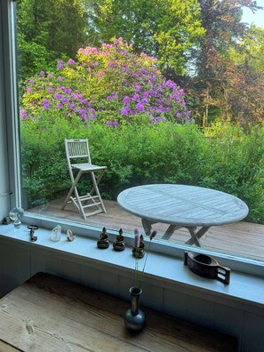 Ein Fenster mit Blick auf einen Garten, ein Holzstuhl, ein runder Tisch und eine Vase mit Pflanzen.