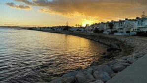 A coastal scene with a row of white buildings along the shore, a rocky breakwater, and the sun setting over the water. - Designer duplex facing the sea in Estepona (Estepona)