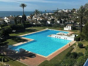 Ein rechteckiger Swimmingpool mit klarem, blauem Wasser, umgeben von einer gepflasterten Liegefläche, mit Blick auf eine Küstenstadt mit Häusern und Palmen.