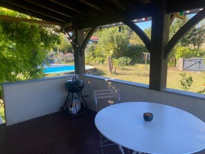 Une terrasse extérieure avec une table ronde, un barbecue et une vue sur une piscine et un jardin.