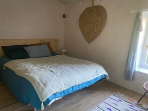 Une chambre à coucher avec une tête de lit en bois, un lit recouvert d’une couvre-lit blanche et bleue, une décoration murale tissée et une fenêtre avec des rideaux.