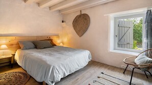 A bedroom with a bed, a window with shutters, a wicker chair, and a hanging heart-shaped decoration. - Le Pigeonnier – Charming Cottage for 2 with Shared Pool (La Tour-Blanche-Cercles)