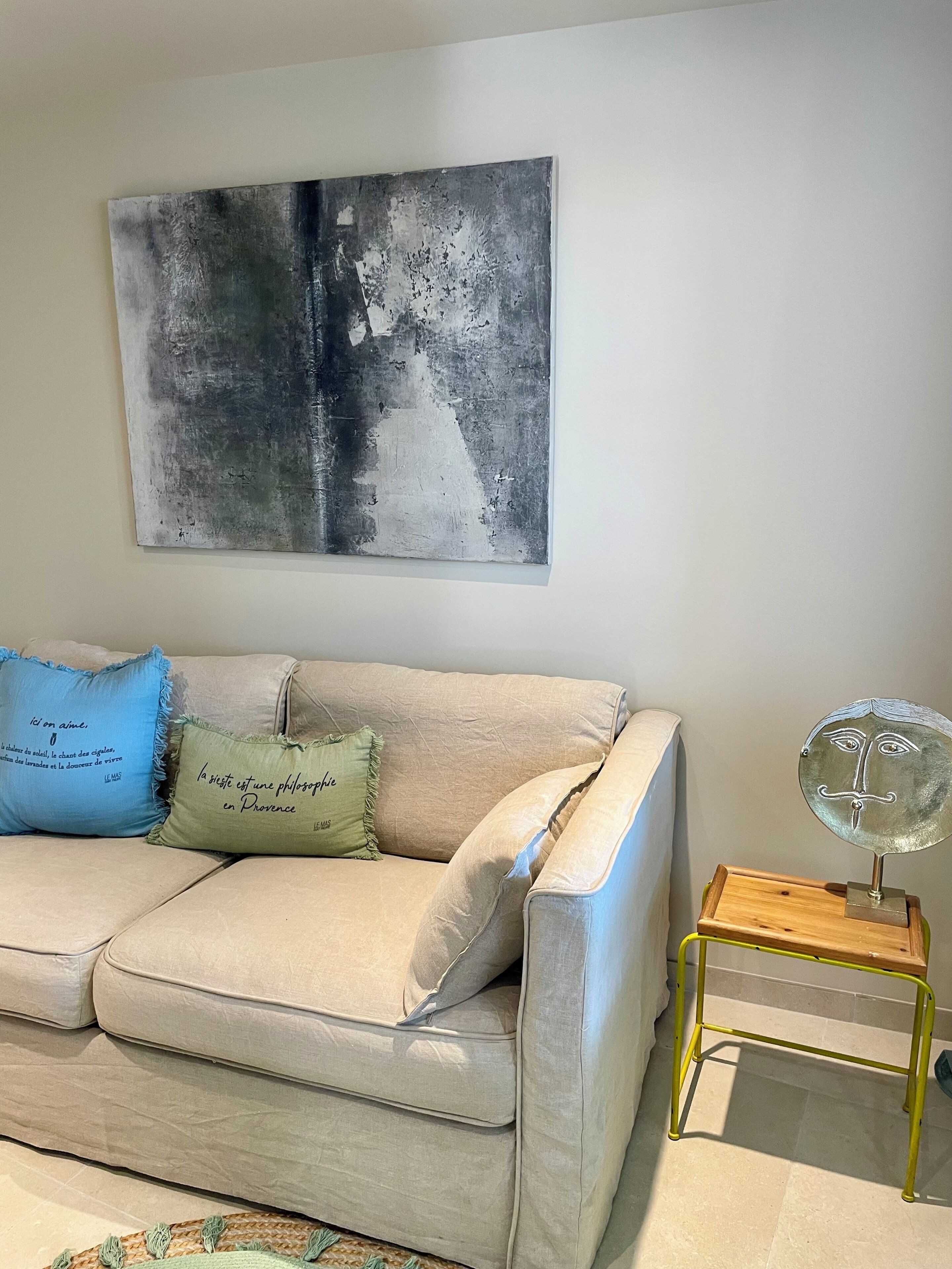 A beige sofa with two pillows, a wall-mounted abstract painting, and a small round table with a metal base.