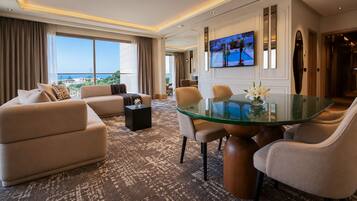 Quarto de hotel moderno com sofá, cadeiras, mesa de vidro, televisão e vista para o mar.