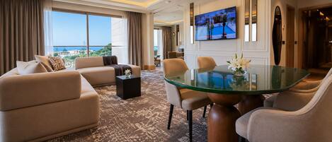 A modern hotel room with a sofa, chairs, a glass table, a TV, and a view of the sea.