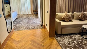 Habitación de hotel con piso de madera, un sofá con cojines estampados, un espejo y vistas a un paisaje urbano por la ventana.