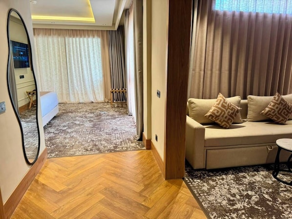 Habitación de hotel con piso de madera, un sofá con cojines estampados, un espejo y vistas a un paisaje urbano por la ventana.