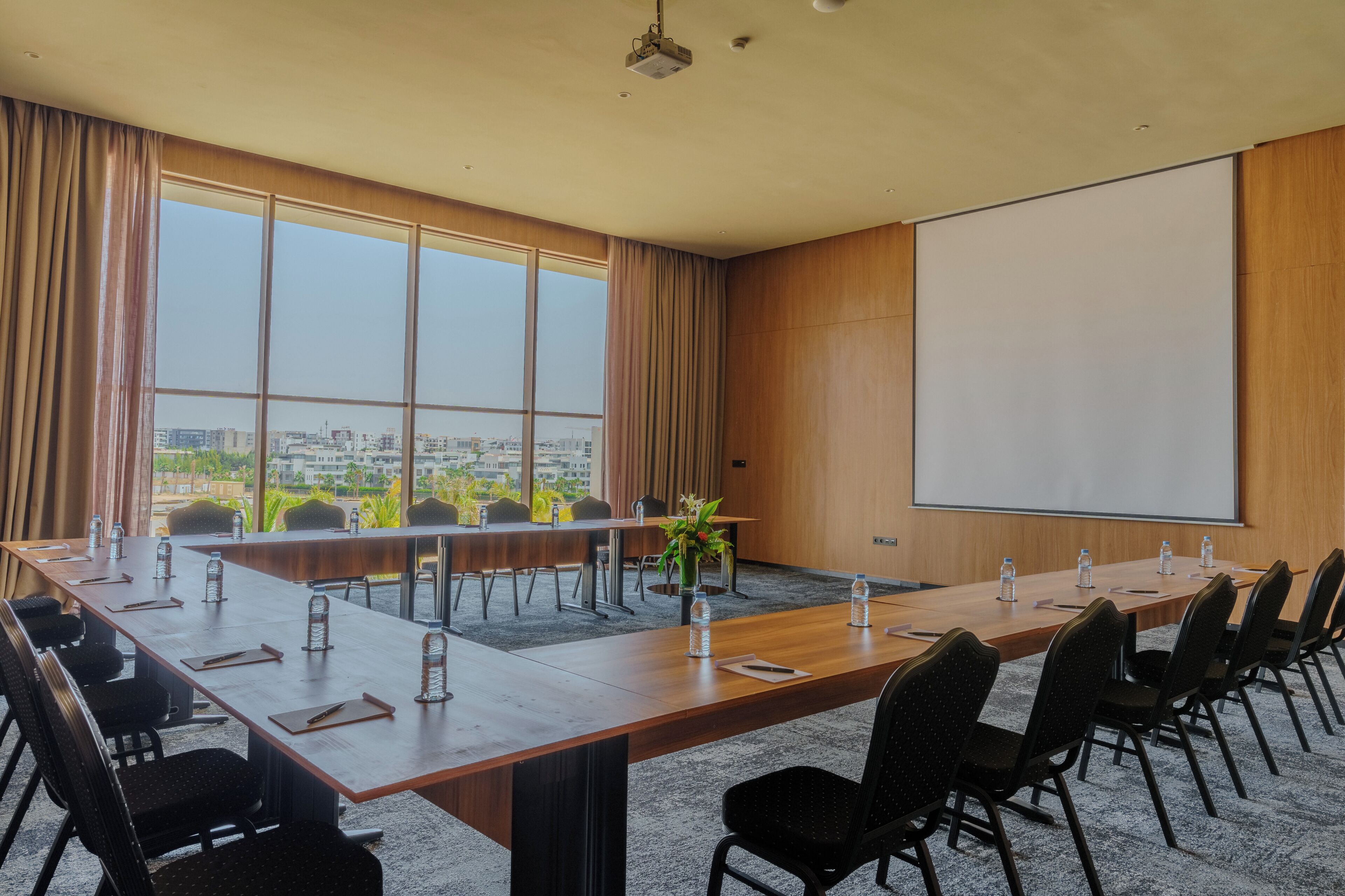 A conference room with a long table, chairs, a projector screen, and a view of the city through large windows.