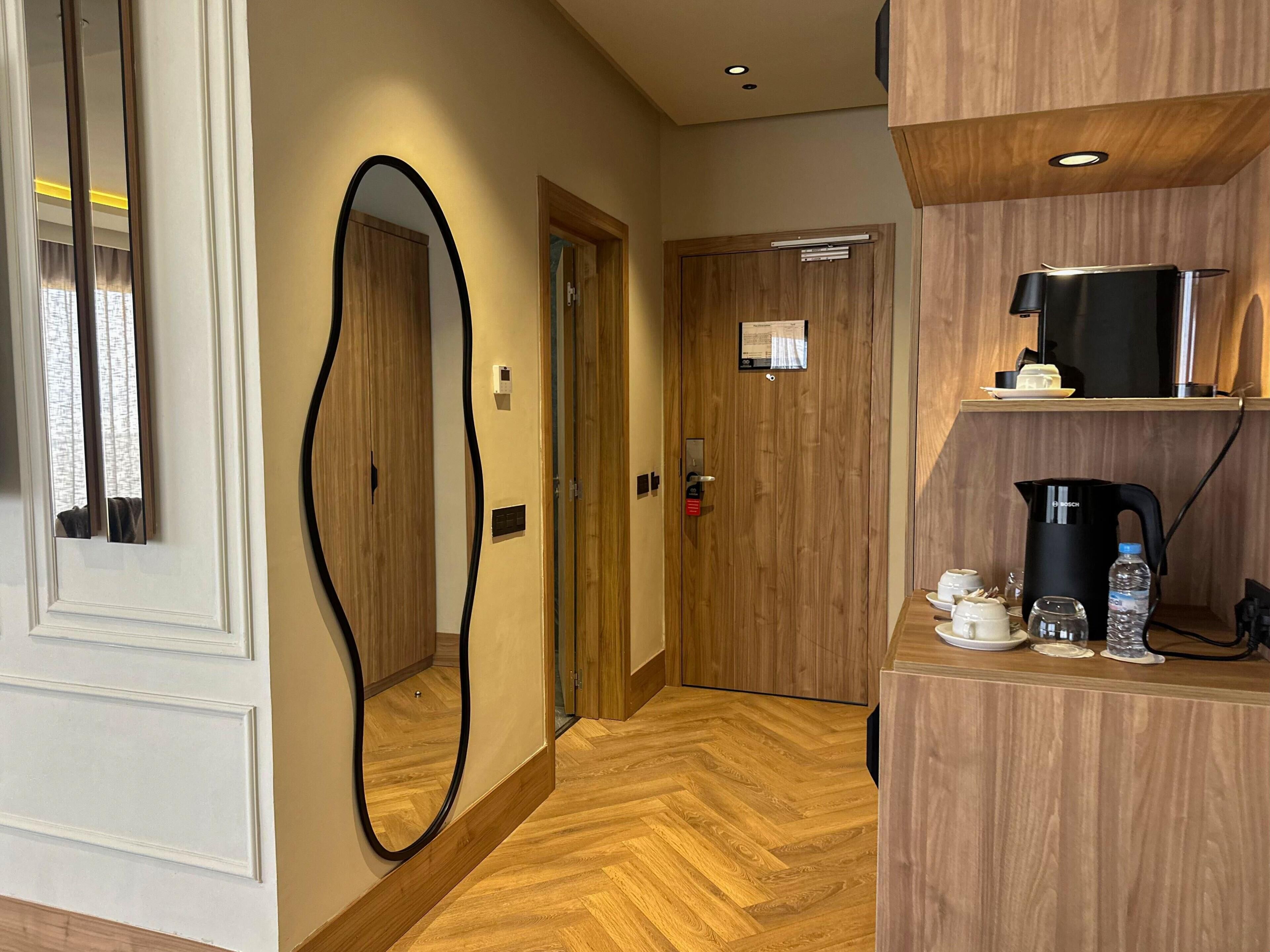 A hotel hallway with a wooden floor, a mirror, a door with a sign, and a coffee station with a kettle and cups.