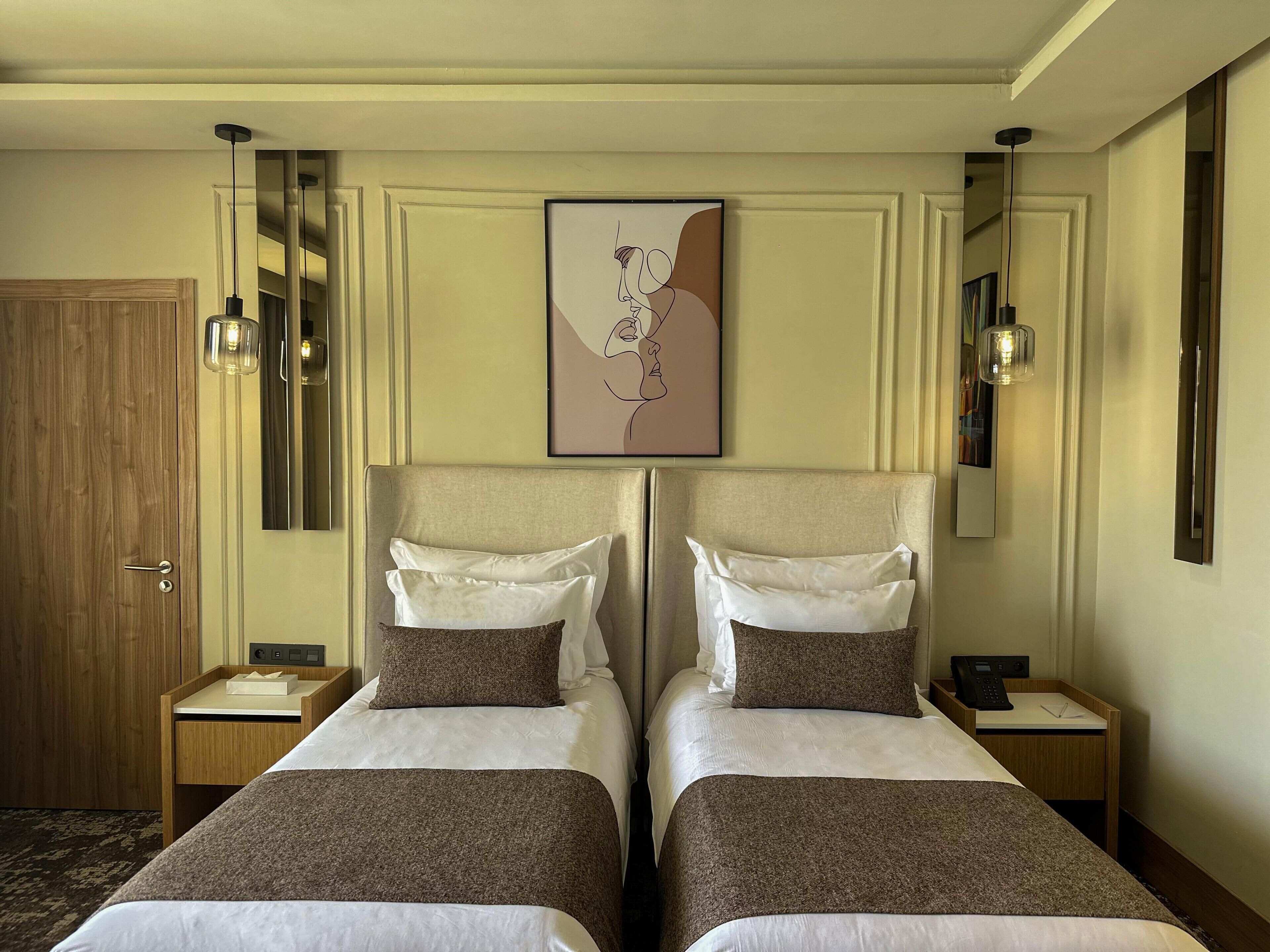 Habitación de hotel con dos camas, una puerta de madera, un cuadro enmarcado en la pared y lámparas de noche.