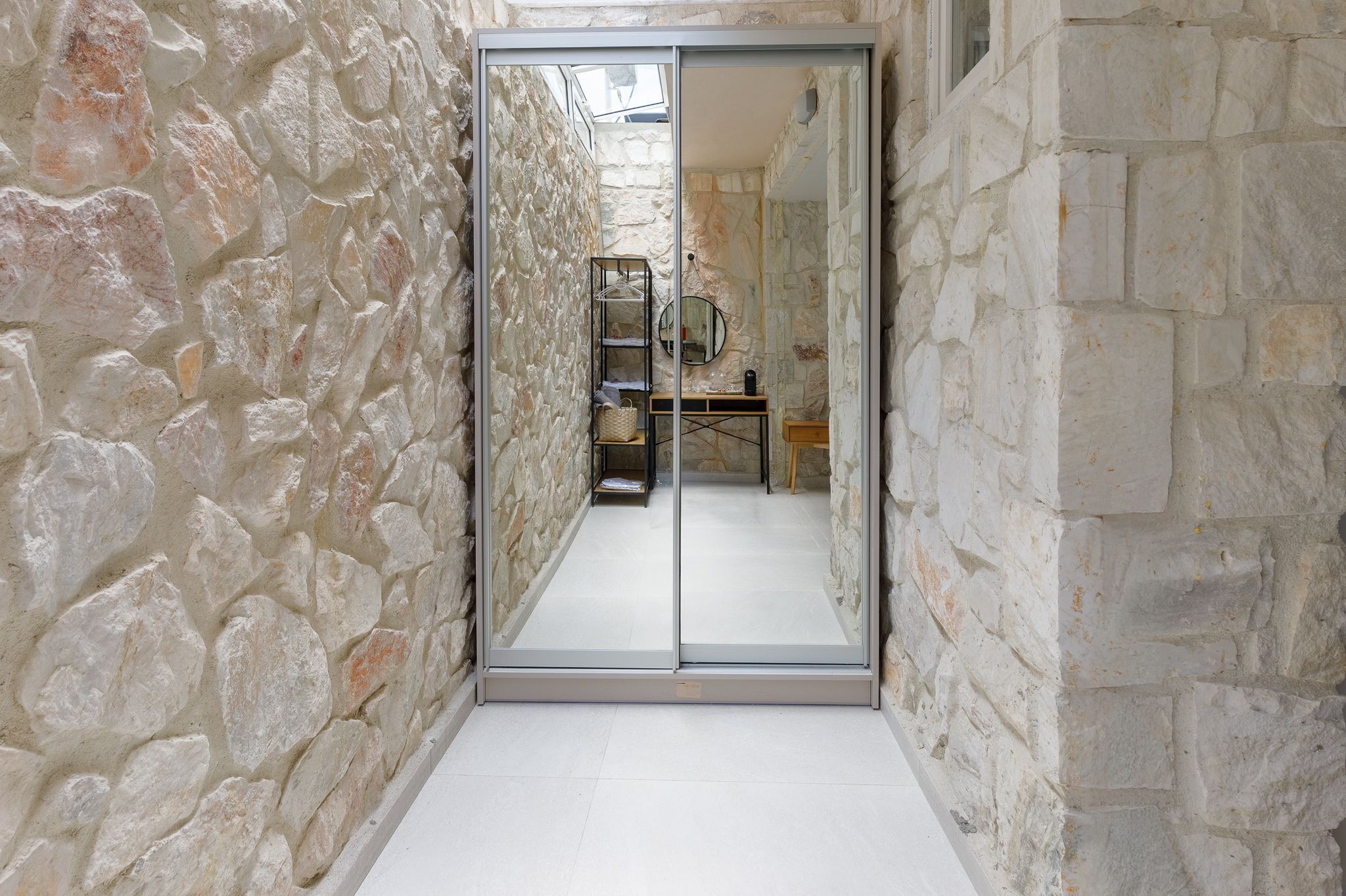A narrow stone corridor with a mirrored door leading to a room with a bed and a small table.