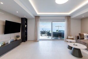 Ein modernes Wohnzimmer mit Flachbildfernseher, einer Couch, zwei Couchtischen und einem Balkon mit Gartenmöbeln.