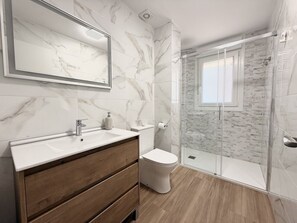 A bathroom with a marble-tiled wall, a wooden vanity, a toilet, and a glass-enclosed shower. - Apartment Los Jazmines 20002 (Santiuste)