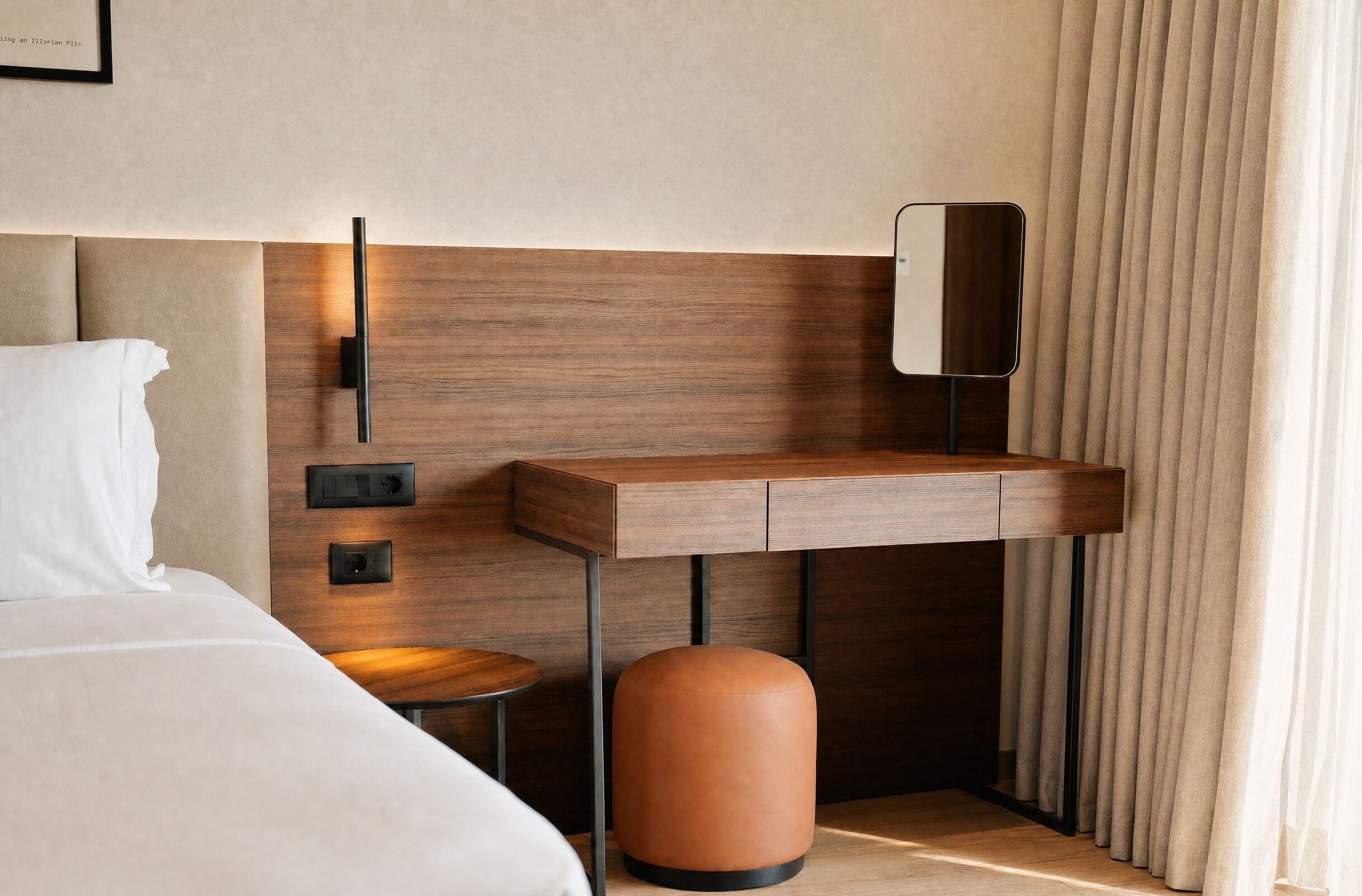 A modern hotel room with a wooden headboard, a bedside table, a small stool, and a round ottoman.