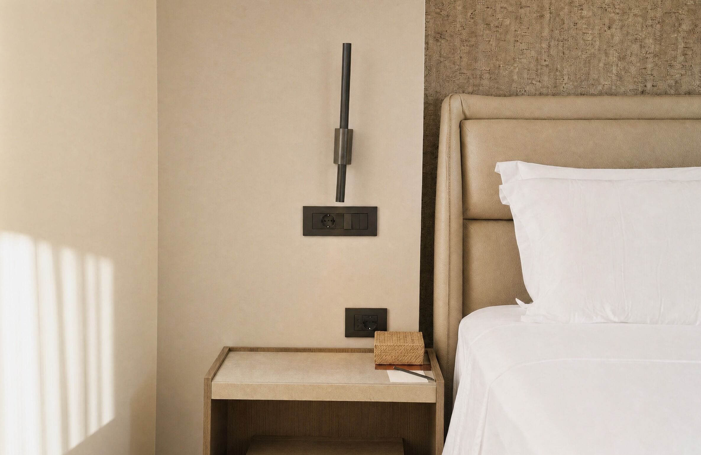 A hotel room with a bed, a nightstand, a wall-mounted light, and a power outlet.