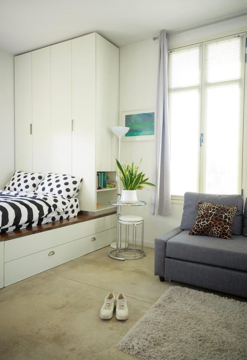 Et soveværelse med en seng, en grå sofa, en hvid garderobe, et lille bord med en plante og et vindue med gardiner.