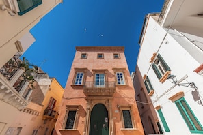 A narrow street with a tall building featuring a green door, balconies, and a clear blue sky.