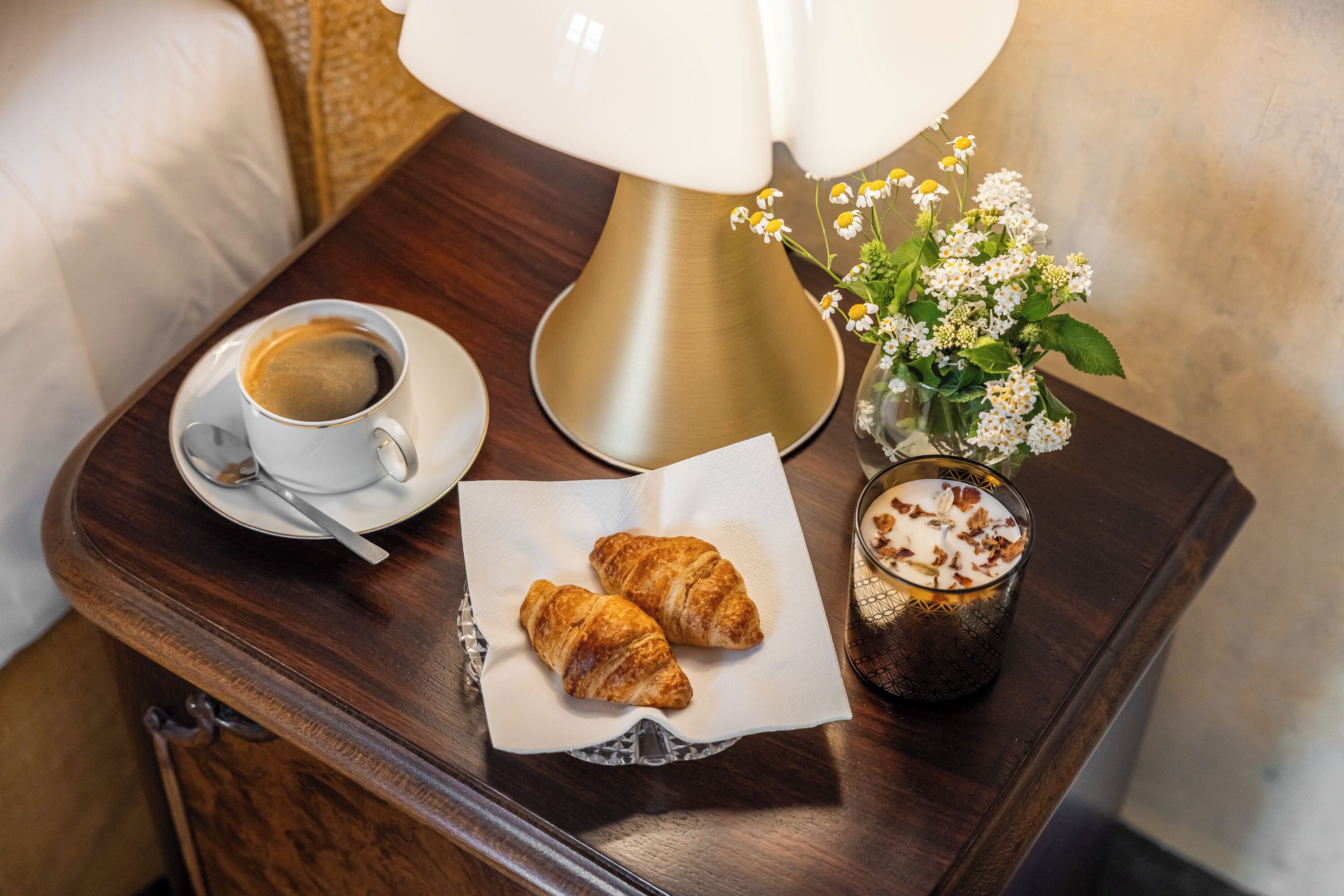 Una mesita de noche de madera con una taza blanca de café, dos croissants, un vaso de leche con virutas de chocolate y un florero con flores blancas.