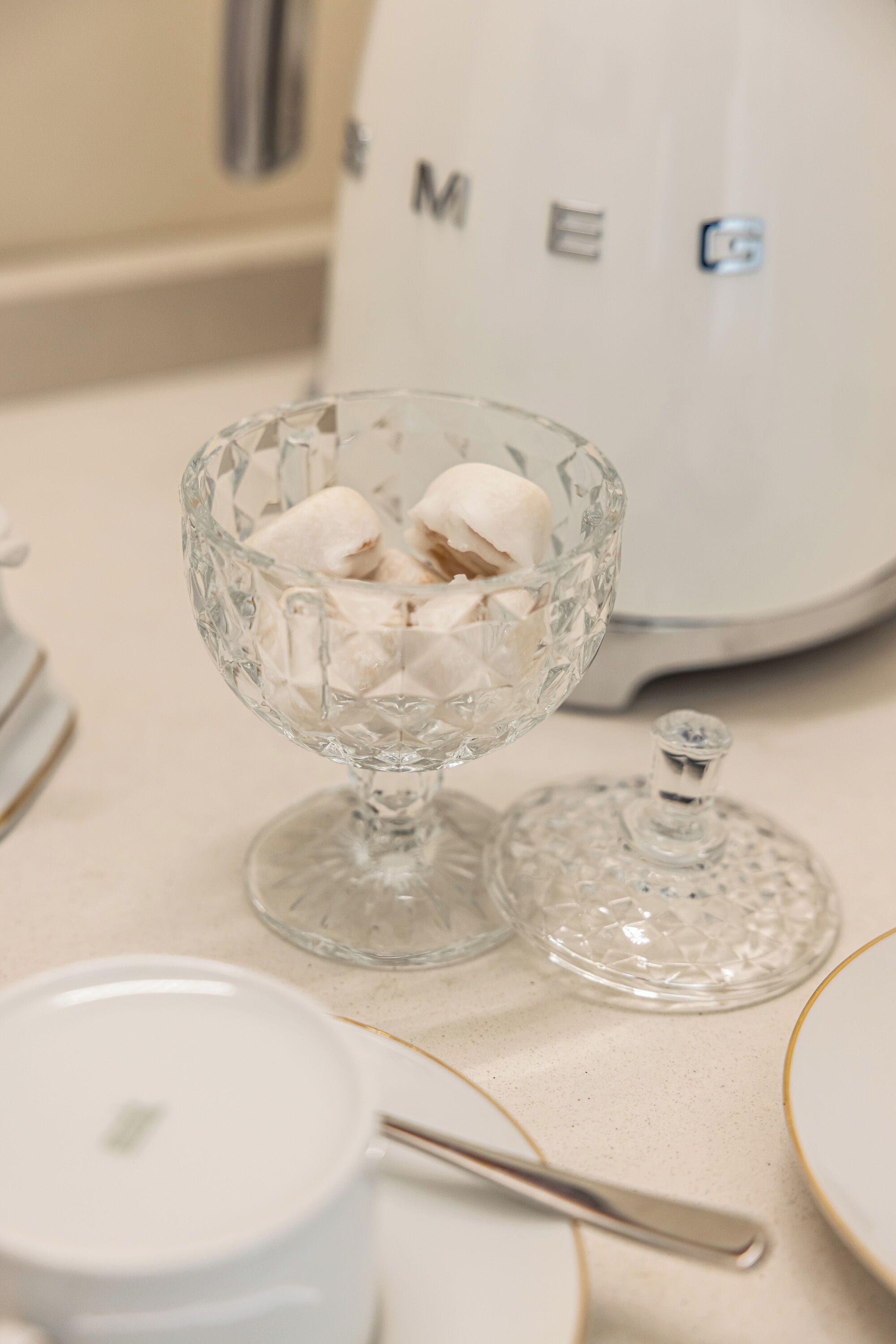 A clear glass bowl with marshmallows on a table, a white appliance in the background, and a spoon.