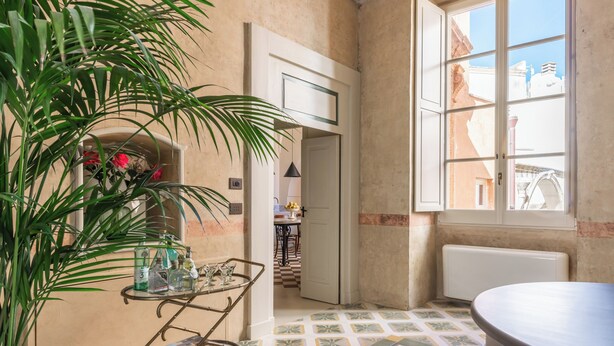 A room with a tiled floor, a potted plant, a metal cart with bottles, a large window, and a door leading to another room.