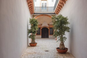 A courtyard with a tiled floor, two potted trees, an arched doorway, and a balcony with a railing.