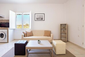 A compact living space with a sofa, a coffee table, a washing machine, and a small shelf. - Santorini's Welcoming Getaway - Blissful & Homely (Pori)