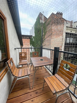 Een balkon met houten meubels, een metalen hekwerk en uitzicht op een bakstenen gebouw.
